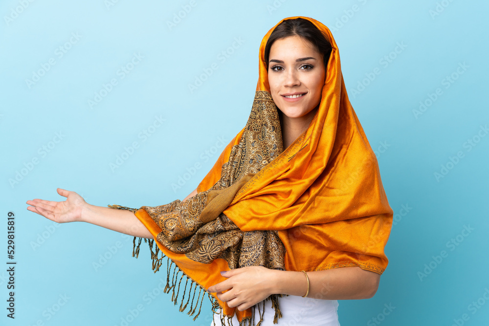 Sticker Young Moroccan woman with traditional costume isolated on blue background extending hands to the side for inviting to come