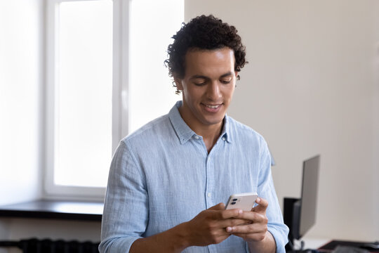 Happy Engaged Latin Office Employee Man Using Online App On Smartphone, Standing At Office Workplace, Reading Text Message, Typing, Chatting On Internet, Smiling, Making Call