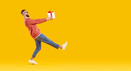 Naklejka premium Happy man carrying a gift box. Side view of a funny young guy in casual clothes holding a present and walking against a bright orange yellow blank empty copy space studio background. Holiday concept