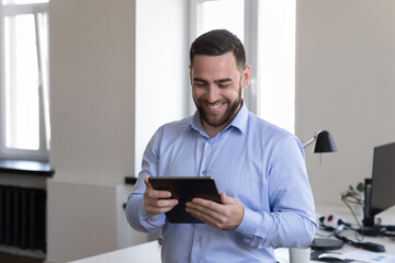 Happy millennial business man watching video content, using online app on tablet, reading good news on gadget screen, laughing, smiling, browsing internet, working in office