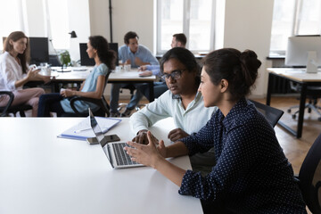 Two serious Indian colleagues talking at work table, sharing laptop in co-working office space,...
