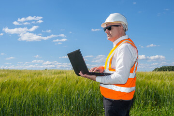 Construction design. Man with laptop in meadow. Design engineer inspects area under construction. Design engineer with laptop is standing in tall grass. Construction bureau worker in protective helmet