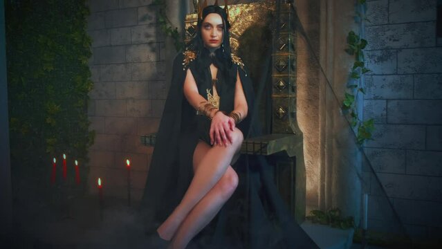 Portrait fantasy gothic woman queen witch sits on throne. Princess girl sexy vampire costume, black dress cape golden crown, long sexy legs pose. Old style room full fog smoke, metal bronze iron chair