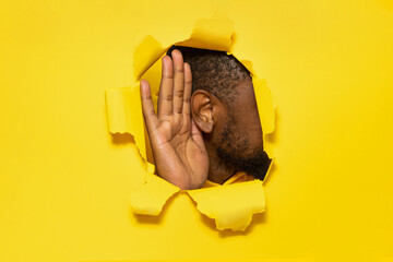 Black man's ear and hand through a torn hole in yellow paper background, free copy space, closeup - obrazy, fototapety, plakaty