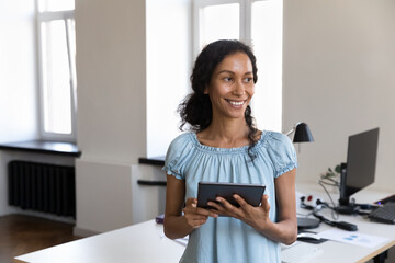 Happy pretty Black employee woman holding tablet computer, standing at office table, looking away, smiling, laughing. African gadget user, businesswoman using digital device, thinking