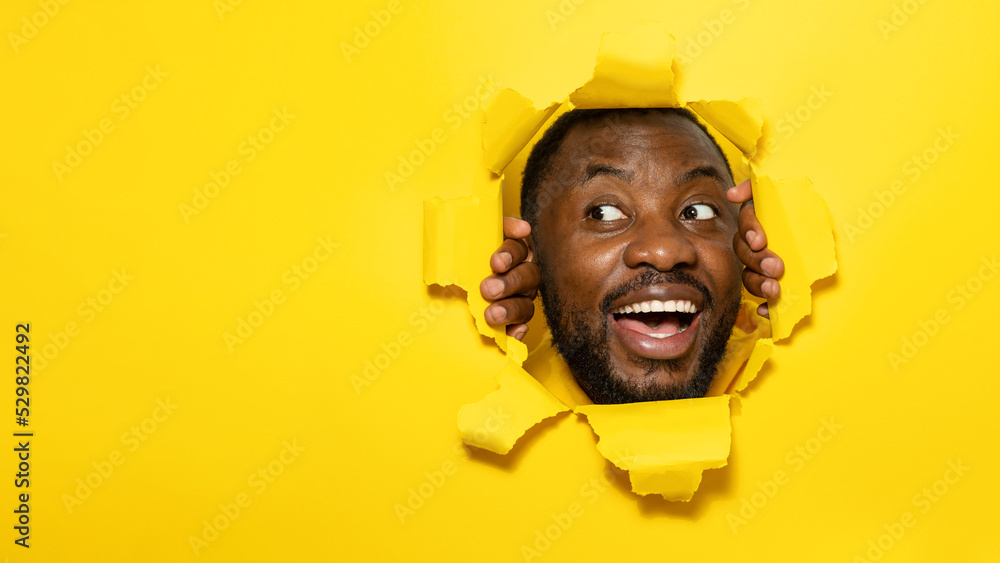 Wall mural excited african american man smiling and looking away while tearing hole in yellow paper background,