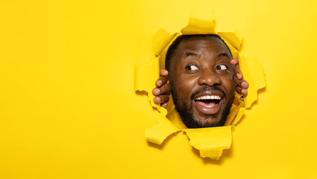 Excited african american man smiling and looking away while tearing hole in yellow paper background, copy space