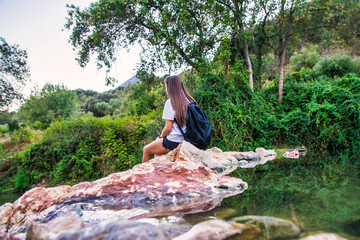 Rear view of a woman sitting on some rocks inside a river and enjoying nature. Nomadic lifestyle. Relaxed young woman in the middle of nature.