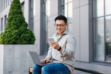Happy asian middle aged businessman texting on cellphone and using laptop, chatting with client outdoors