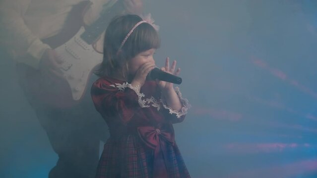Little girl on stage in vintage dress, she sings into microphone and dances, her father plays an electric guitar. Color music is shining and smoke is billowing. Family time, creativity and hobbies