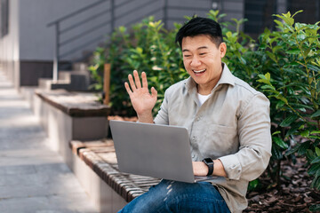 Cheerful asian businessman looking at laptop webcam and waving hand, greeting client, sitting near office building