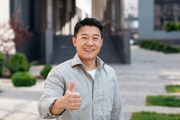 Portrait of positive asian man standing outdoors and gesturing thumb up, walking in urban city...