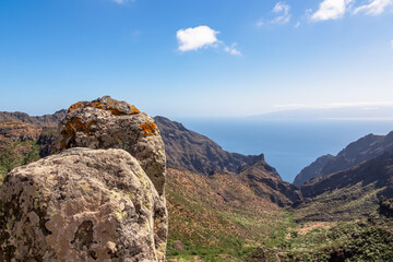 Aerial panoramic view on the Teno mountain massif near Masca, Tenerife, Canary Islands, Spain, Europe. Island La Gomera in the distance. Massive rock with orange collared lichen in focused foreground