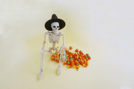 Skeleton WIth Black Pointy Whitch Hat Sitting with Orange Candy Corn on Yellow Background for Halloween
