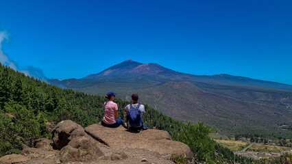 Fototapeta premium Sitting couple with scenic view on volcano Pico del Teide surrounded by pine tree forest, Teno mountain, Tenerife, Canary Islands, Spain, Europe. Hiking trail from Santiago to Masca via Pico Verde