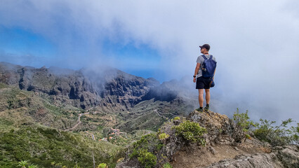 Naklejka na ściany i meble Man with backpack enjoying view on Teno mountain range near Masca village, Tenerife, Canary Islands, Spain, Europe. Fog going down the valley creates a mystical atmosphere. Remote hiking trail