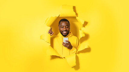 Happy african american man using cellphone and smiling, standing in torn paper hole, panorama