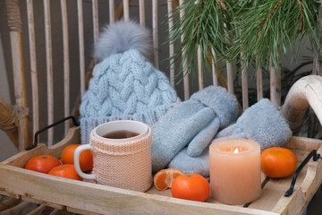Obraz na płótnie Canvas cozy woolen things, a hat and mittens, a mug of hot tea on a tray, delicious vitamin tangerines, a candle is burning, a wicker garden chair, a concept vacation, a weekend in nature
