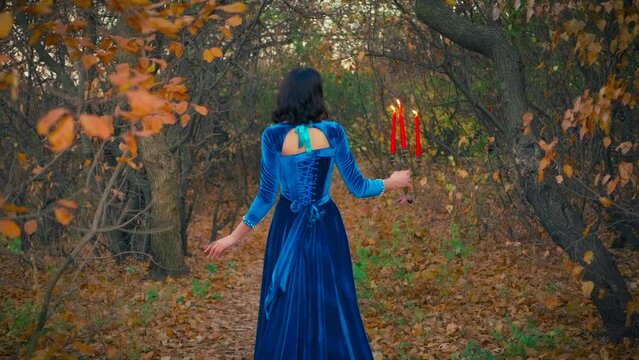 Beauty autumn forest bare black tree trunks. Gothic elegant fantasy woman queen. girl vampire princess turned away, holding candlestick red candles burning fire. Long blue vintage dress back rear view