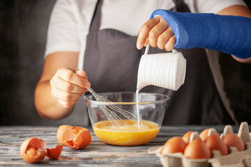 Woman with a broken hand in a blue cast cooks raw eggs in the kitchen.