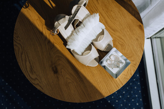 Wedding details, accessories close-up on a wooden table in the morning: bride's shoes, rings, jewelry box, garter. Photo, top view.