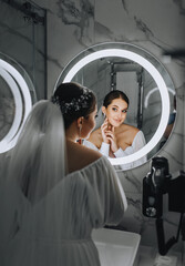 A beautiful, young bride model stands in front of a round mirror with a lamp in the bathroom,...