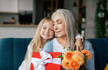 Glad caucasian little granddaughter hugs senior woman, gives flowers, box with gift congratulates, have fun