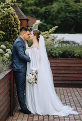 A stylish, young groom in a blue suit and a beautiful bride in a white dress are hugging while standing in a park in nature. Wedding photography, portrait of smiling newlyweds.