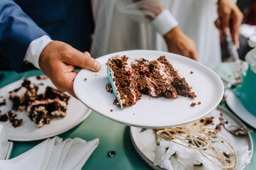 The bride and groom are cutting with a knife a sweet, delicious, sweet wedding cake standing on the table into large pieces close-up holding a plate in their hands at the ceremony.