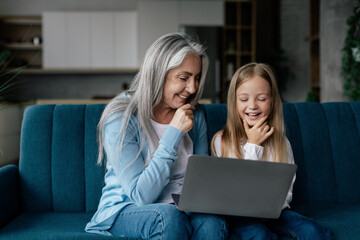 Cheerful caucasian little granddaughter and senior grandma think, watch video on computer, study...