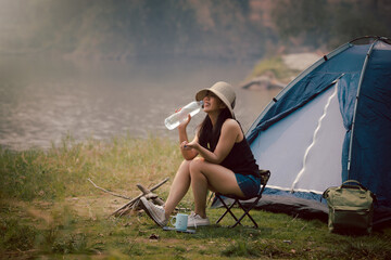 Happy woman drinking water and sitting on the camping chair in front of the camping tent at meadow...