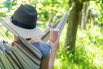 Naklejka na ściany i meble Young woman with hat resting in comfortable hammock at green garden with Glass of fresh strawberry milkshake, smoothie. Healthy food and drink concept.