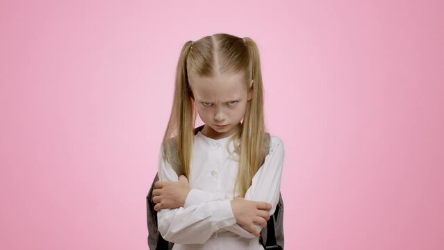 Victim of bullying. Little grumpy schoolgirl with backpack posing with crossed arms and looking sullenly to camera