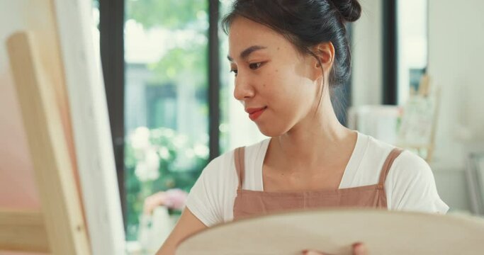 Happy youth Asia Female wear apron holding palette color use brush painting on canvas create artwork in cozy workshop at home. Contemporary Painter Abstract Modern Art, Creativity and people concept.