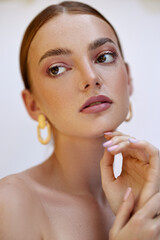 Female model with red hair, brown eyes, freckled skin, poses for a beauty shoot. She wears gold...