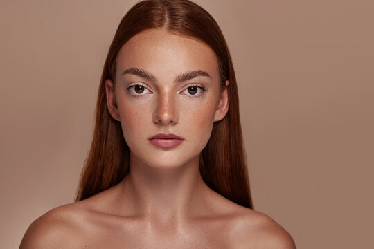 Beauty photoshoot.  Female model with red hair, brown eyes, freckles, clear skin, against a beige / brown backdrop. 