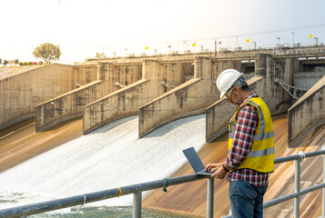 Aืn Engineer standing by the dam. He is wearing a white hard hat and yellow transparent vest.
