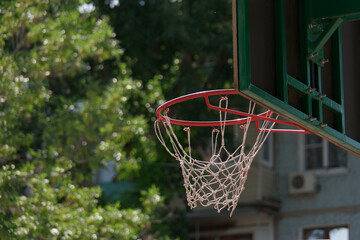 Rear view of red metal basketball hoop with white net and backboard against the green park trees and nearby building
