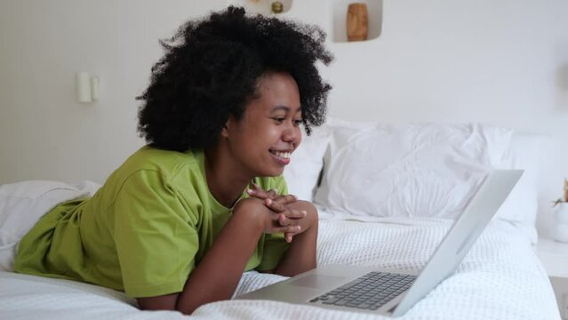 Happy Afro Asian woman on bed chatting with friends via video call on laptop from her bedroom. Black woman with afra hairstyle is happy that she can chat with friends online. Life style concept.