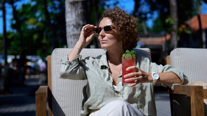 A beautiful woman in sunglasses on vacation enjoys basking in the sun while drinking a soft drink with mint. A woman on vacation relaxes from the routine. Curly woman is resting.
