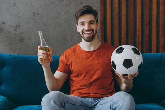 Young fan man in red t-shirt cheer up support football team sport team hold soccer ball watch tv live stream sit on blue sofa stay at home flat rest relax spend free spare time in living room indoor