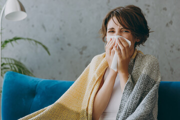 Young sick ill sad woman wear white wrapped in plaid sneezing blow nose use napkin sit on blue sofa couch stay at home hotel flat spend time in living room indoors grey wall People healthcare concept