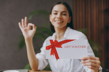 Young happy latin woman in white shirt hold gift certificate coupon voucher card show ok sit at table in coffee shop cafe restaurant indoors. Freelance mobile office business concept. Focus on flyer - obrazy, fototapety, plakaty