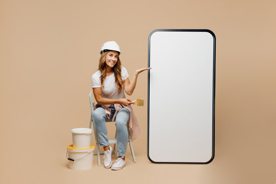 Full size young laborer handyman woman in white tshirt helmet point blank screen area mobile cell phone hold brush paint isolated on plain beige background Instruments accessories for renovation room