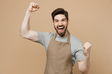 Young happy fun overjoyed excited man barista barman employee wears brown apron work in coffee shop do winner gesture isolated on plain pastel light beige background. Small business startup concept.