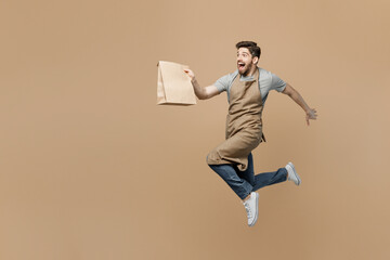 Full body fun young man barista barman employee wear brown apron work in coffee shop run jump hold blank craft paper bag isolated on plain pastel light beige background Small business startup concept