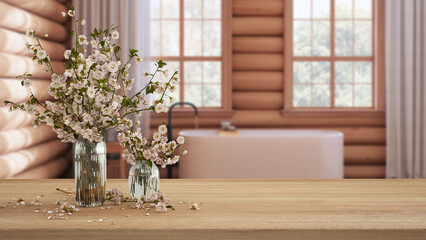 Wooden table, desk or shelf close up with branches of cherry blossoms in glass vase over blurred view of log cabin bathroom with bathtub, rustic interior design concept