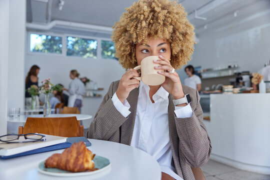 Elegant woman in stylish outfit drinks aromatic coffee eats croissant poses in cozy cafeteria has lunch break enjoys leisure time in public place focused somewhere thoughtfully. People and lifestyle