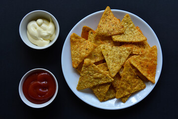Delicious peppery nachos on a plate with sauces on a black background. Delicious snack with corn nachos close-up.
