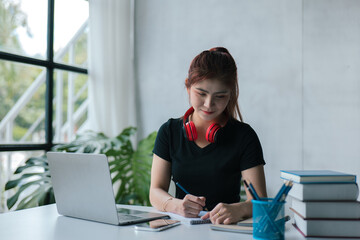 Young beautiful woman is studying online via laptop and writing on note book at home.